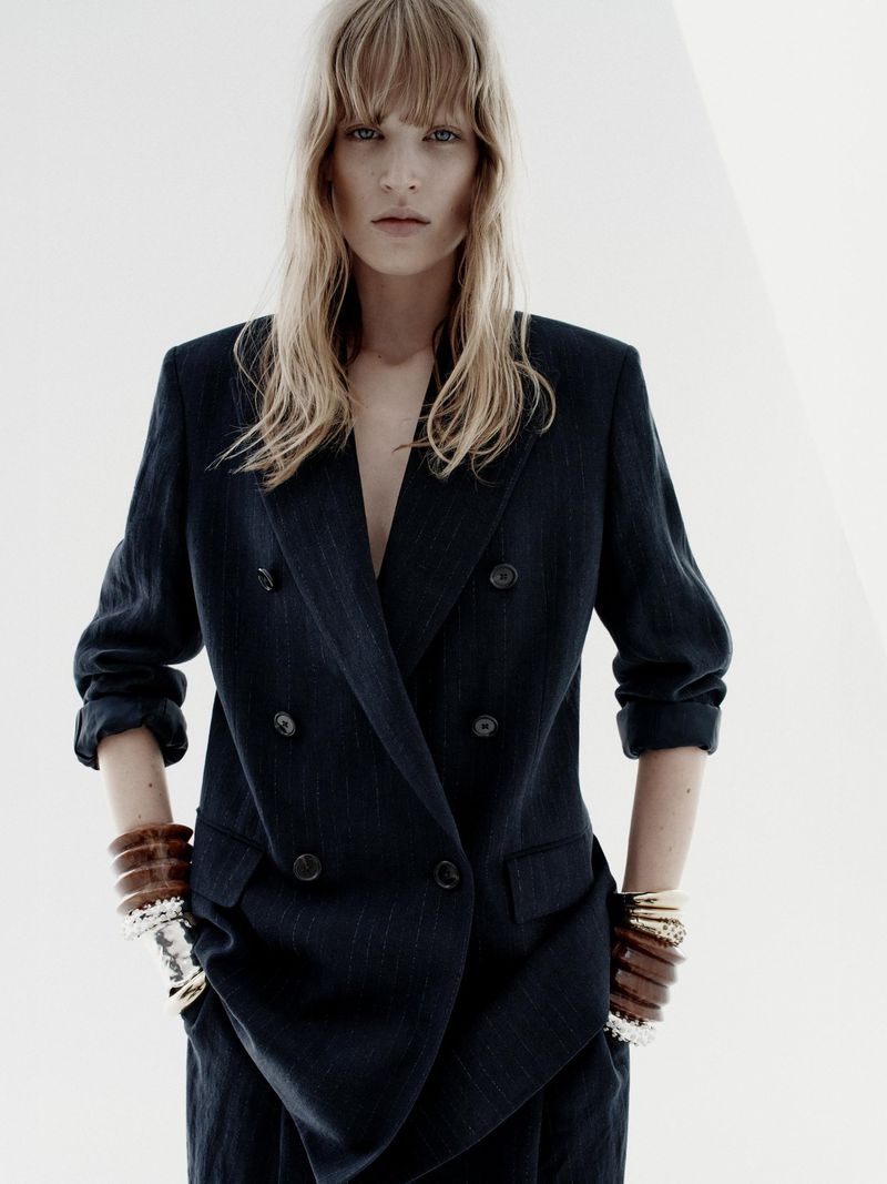Indoor shot of a woman wearing a navy blue double-breasted blazer with bracelets on both wrists. She has light blonde hair.