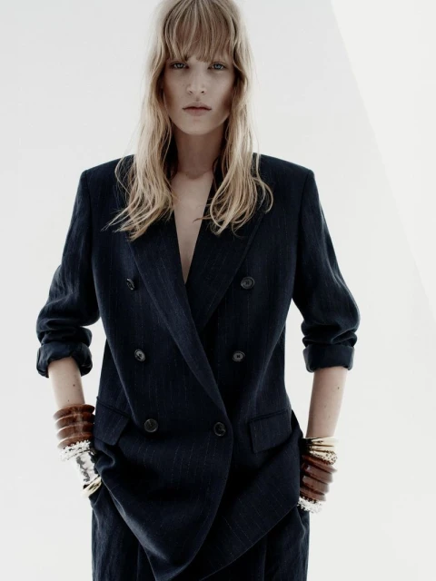Indoor shot of a woman wearing a navy blue double-breasted blazer with bracelets on both wrists. She has light blonde hair.