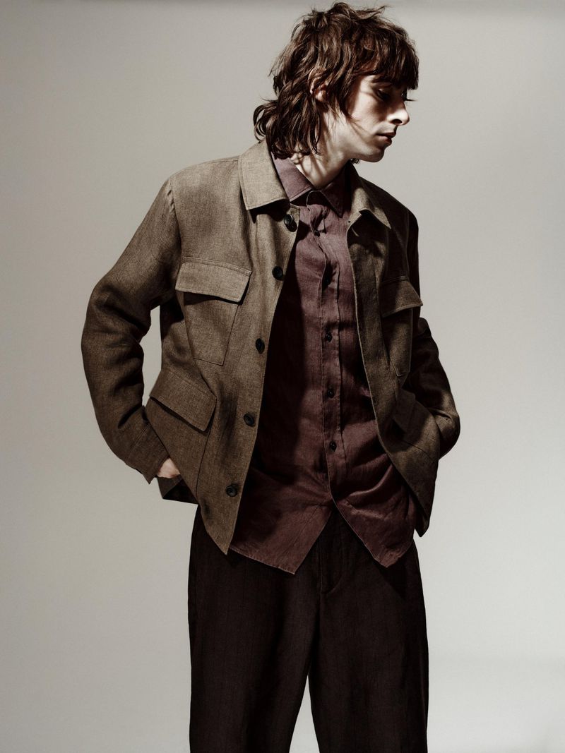 A young man poses in brown tones, wearing a brown jacket over a burgundy shirt and dark brown trousers, in a studio setting.