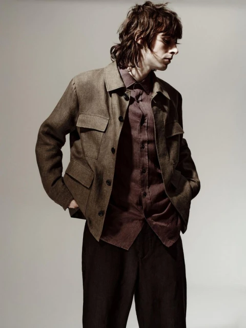 A young man poses in brown tones, wearing a brown jacket over a burgundy shirt and dark brown trousers, in a studio setting.