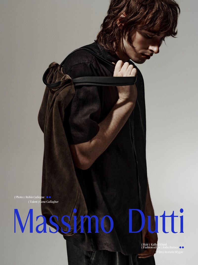 Fashion shot of a male model with shoulder-length wavy hair wearing a dark shirt with a tan jacket draped over his shoulder.
