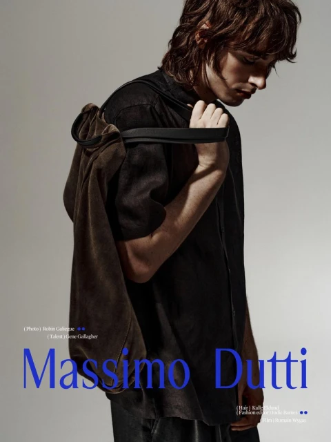 Fashion shot of a male model with shoulder-length wavy hair wearing a dark shirt with a tan jacket draped over his shoulder.