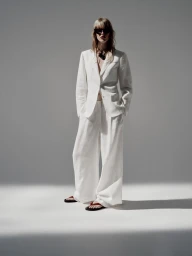 Model wearing a white suit and sunglasses standing against a light grey seamless studio background.