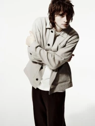 Young man with dark hair in a light-coloured collared jacket and dark trousers standing against a plain white studio background.