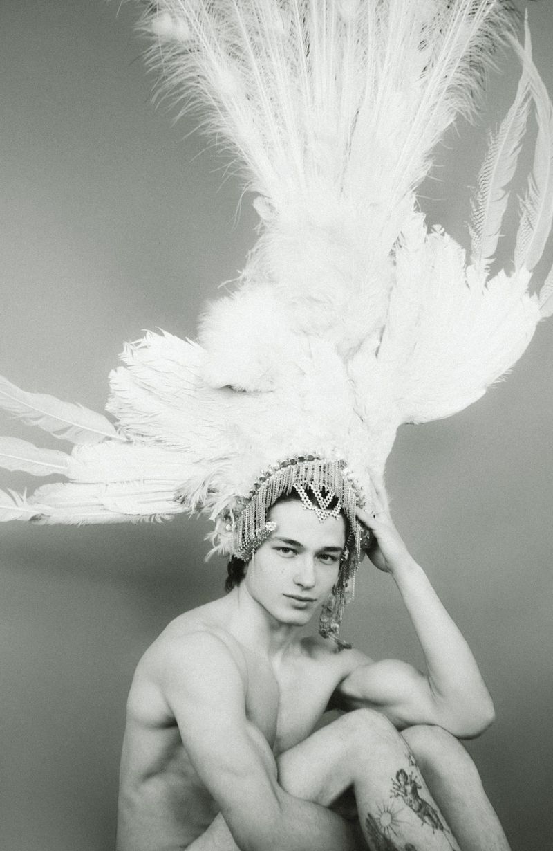 Portrait of a shirtless man wearing a feathered headdress with jeweled detailing, looking at the camera against a grey background.