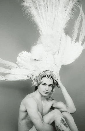 Portrait of a shirtless man wearing a feathered headdress with jeweled detailing, looking at the camera against a grey background.