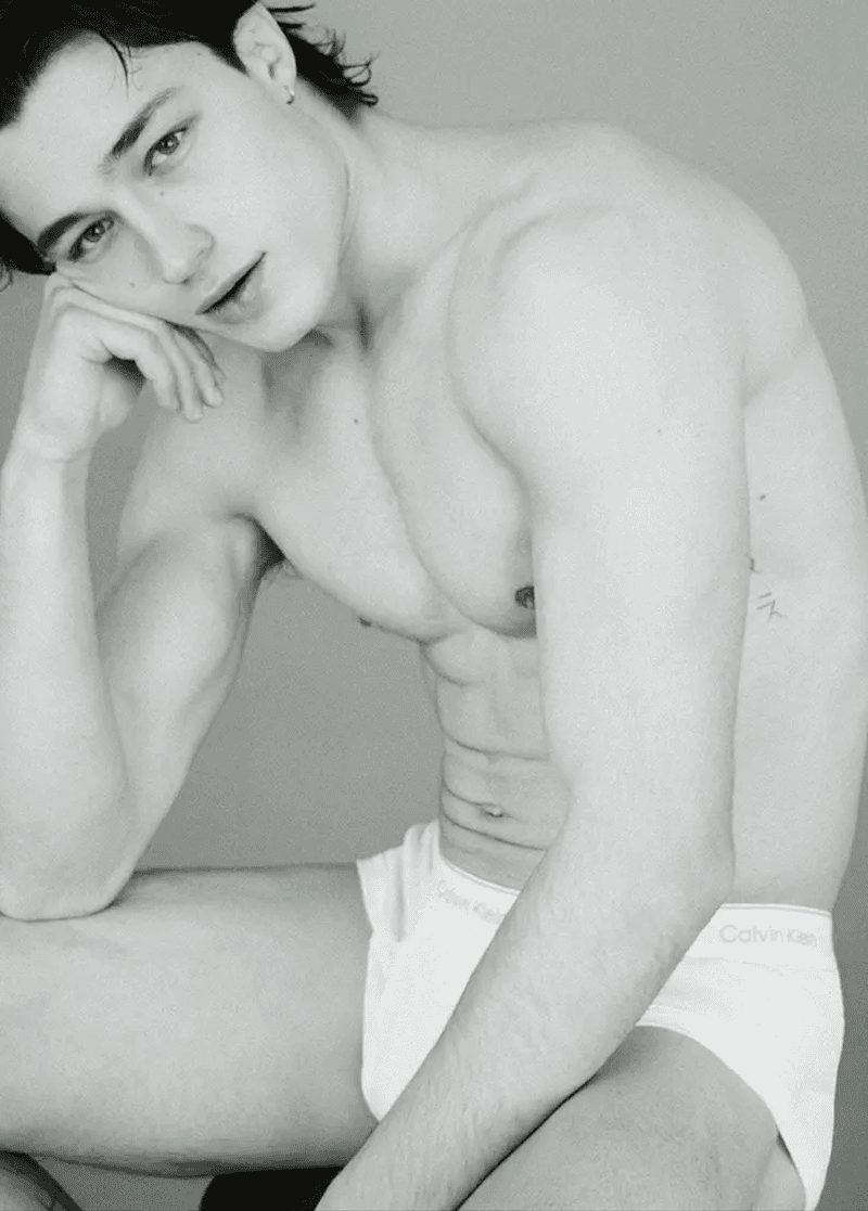 Black-and-white close-up of a shirtless young adult male model in underwear, seated indoors with hand on head, looking at camera.