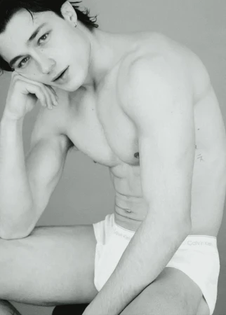 Black-and-white close-up of a shirtless young adult male model in underwear, seated indoors with hand on head, looking at camera.