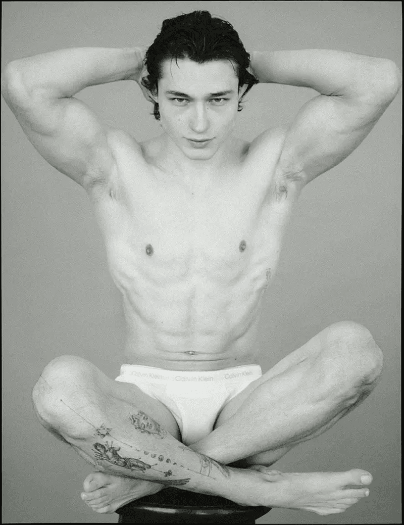 Black and white portrait of a shirtless adult male, sitting cross-legged with hands behind his head, showcasing a slim, muscular build and tattoo.