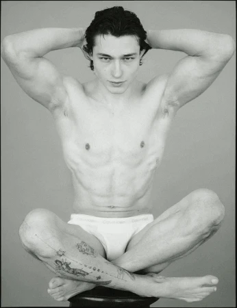 Black and white portrait of a shirtless adult male, sitting cross-legged with hands behind his head, showcasing a slim, muscular build and tattoo.