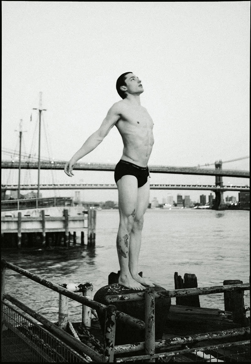 Monochrome photograph of a shirtless man standing atop a weathered, metal structure with a waterfront and city skyline in the backdrop.
