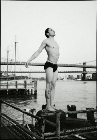 Monochrome photograph of a shirtless man standing atop a weathered, metal structure with a waterfront and city skyline in the backdrop.