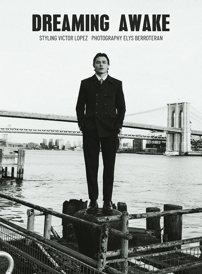 A man in a suit stands on a pier in a black-and-white fashion shot, with the Brooklyn Bridge and city skyline in the background.
