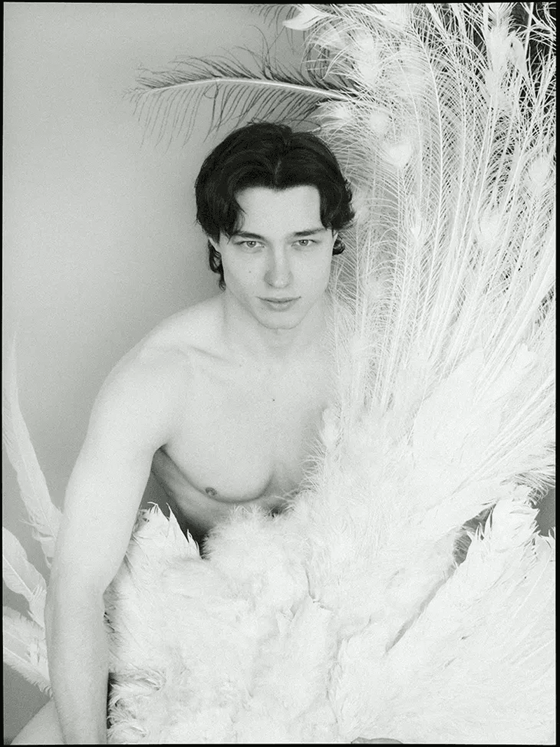 Monochrome portrait of a young adult man with dark hair partially obscured by white peacock feathers in a photo studio setting.