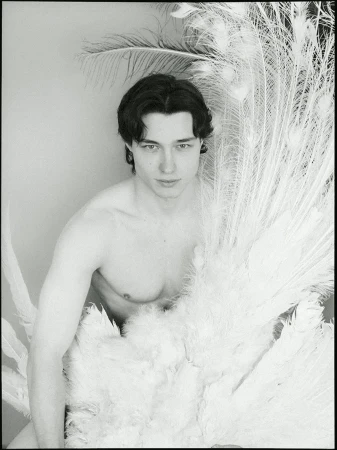 Monochrome portrait of a young adult man with dark hair partially obscured by white peacock feathers in a photo studio setting.