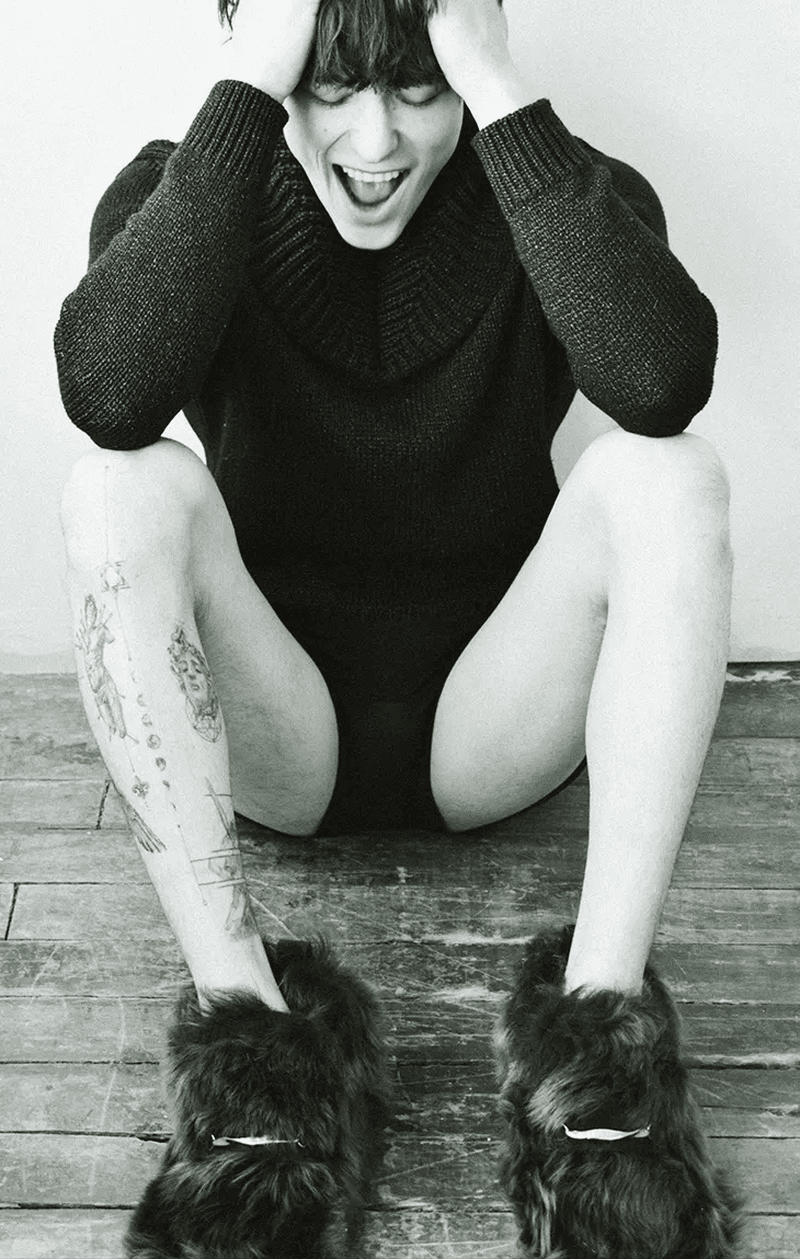Monochromatic fashion portrait of a person in a dark sweater, sitting on the floor with fur slippers and tattoos.
