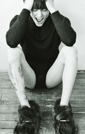 Monochromatic fashion portrait of a person in a dark sweater, sitting on the floor with fur slippers and tattoos.