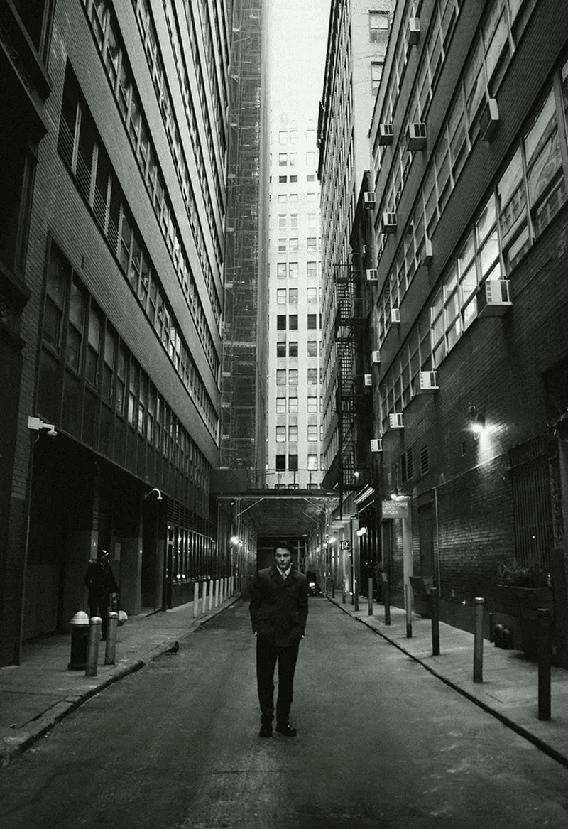 Monochrome portrait of a man in a dark coat standing in an urban alleyway lined with tall buildings and architectural details.