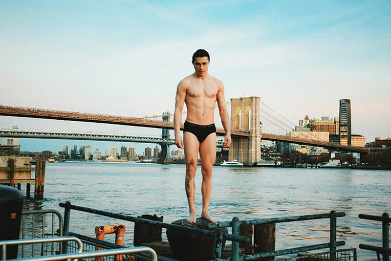 Athletic adult male stands on a pier with a city skyline and bridge visible in the background, shot in eye-level medium framing.