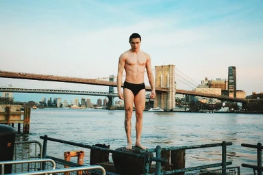 Shirtless man wearing black briefs standing on a pier in front of a bridge and river skyline