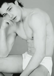 Monochrome portrait of a shirtless man wearing white briefs, posing with his hand resting under his chin against a plain studio background.