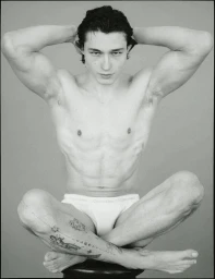 Shirtless man with tattoos sitting cross-legged wearing white underwear against a seamless grey backdrop.