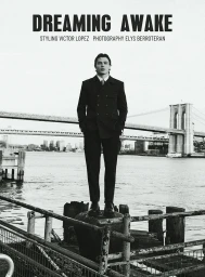 A man in a dark suit standing on a pier with a suspension bridge in the background