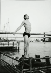 Man standing shirtless wearing dark briefs on a waterfront pier with suspension bridges in the background.