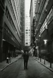 Man standing in the center of a narrow city alleyway framed by tall brick buildings under high contrast monochrome lighting