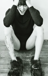 Monochrome portrait of a seated man wearing a dark turtleneck sweater and fuzzy fur slippers, holding his head with both hands.
