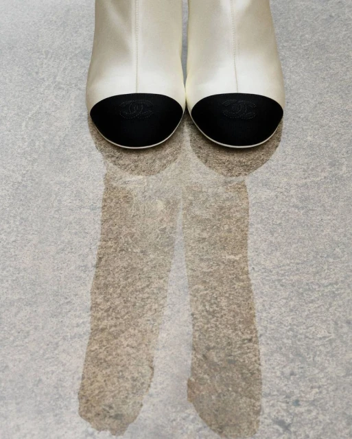 Close-up studio shot of a pair of white ankle boots with black cap toes, and reflections on a mottled grey surface.