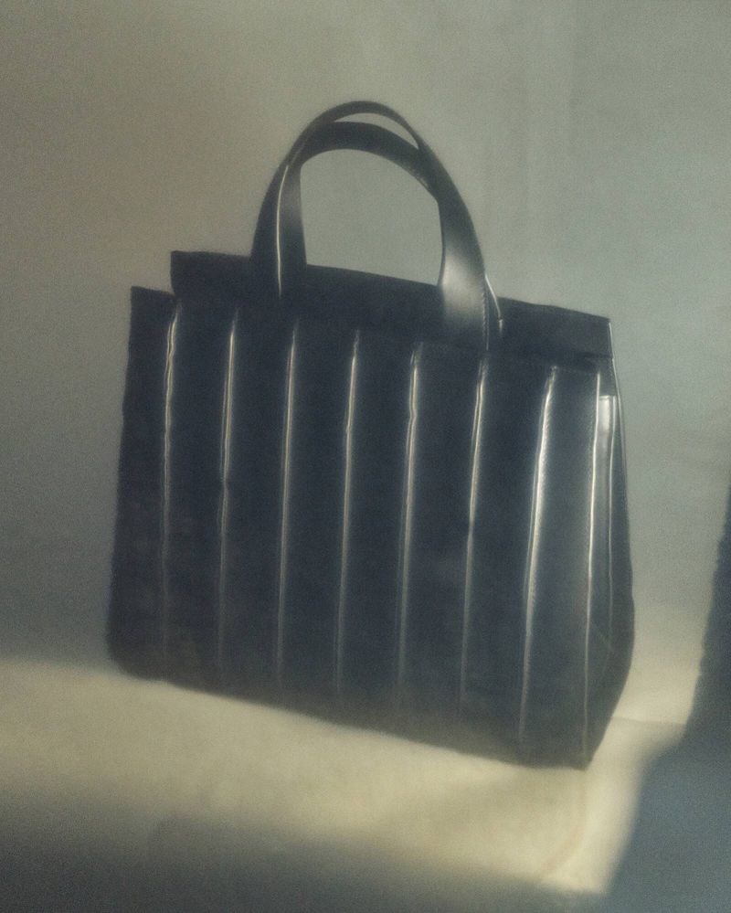 Elegant black leather tote bag with structured vertical panels and rounded handles, resting on a soft, gradient surface.