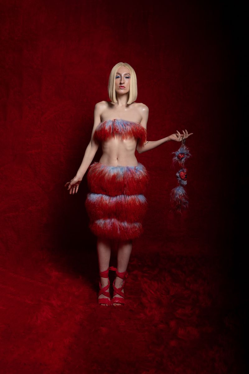 Full shot of a fashion model posing in a tiered, fringed dress against a textured red background with dramatic lighting.