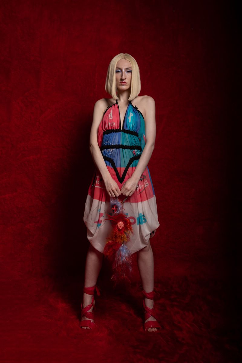 Woman with blonde bob wearing colorful dress and red heels poses against a red background in a fashion photography studio shot.
