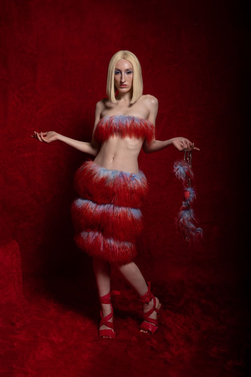 Woman in avant-garde fashion posing against a red backdrop, featuring a bustier and skirt made of red and blue faux fur.