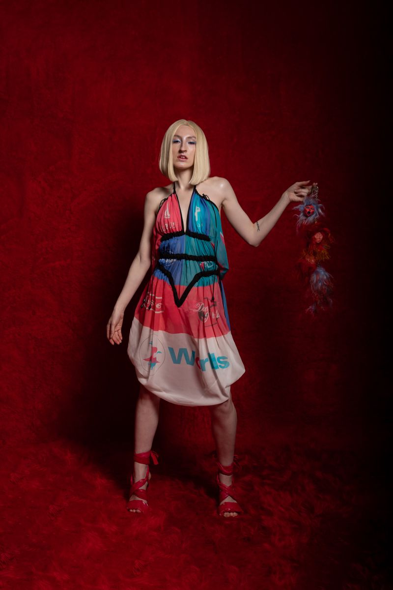 A woman with blonde hair poses in a halter dress against a red background. She holds a feathered object in her right hand.