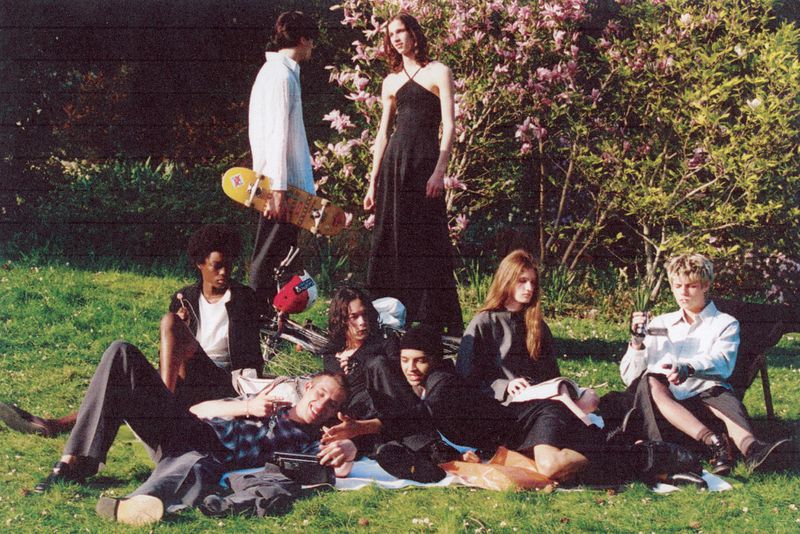 Group of young adults gathers on a grassy lawn in a park-like setting with blooming trees, some sitting on a blanket, others on chairs.