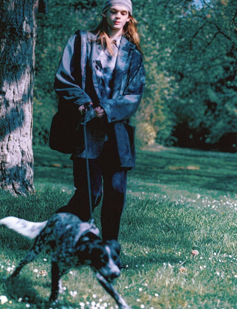 A person walks a speckled dog on a leash in a sunny park setting. The person wears a beanie, patterned jacket, and dark pants.