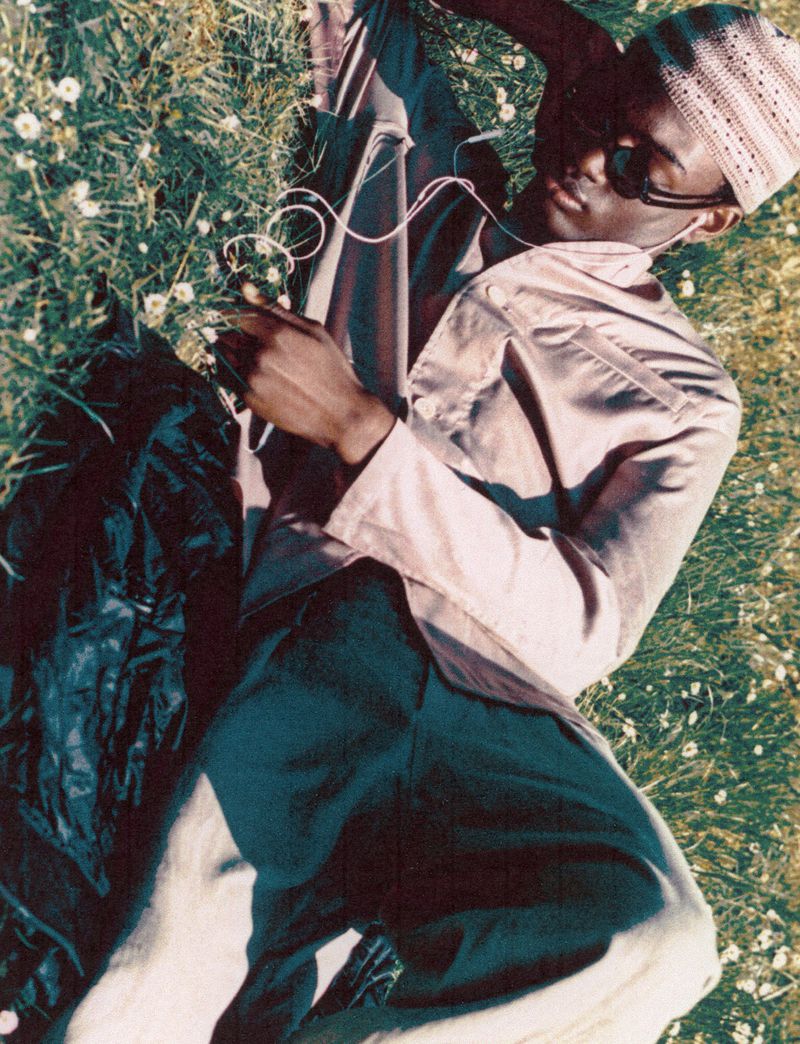 Young man with sunglasses and hat lying on grass with daisies listening to headphones, wearing a light-colored jacket and dark pants.