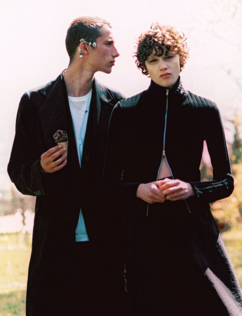 Two stylish adults in a blurred outdoor setting: one holds an ice cream cone, both wear dark clothing, and the photo has a vintage tone.