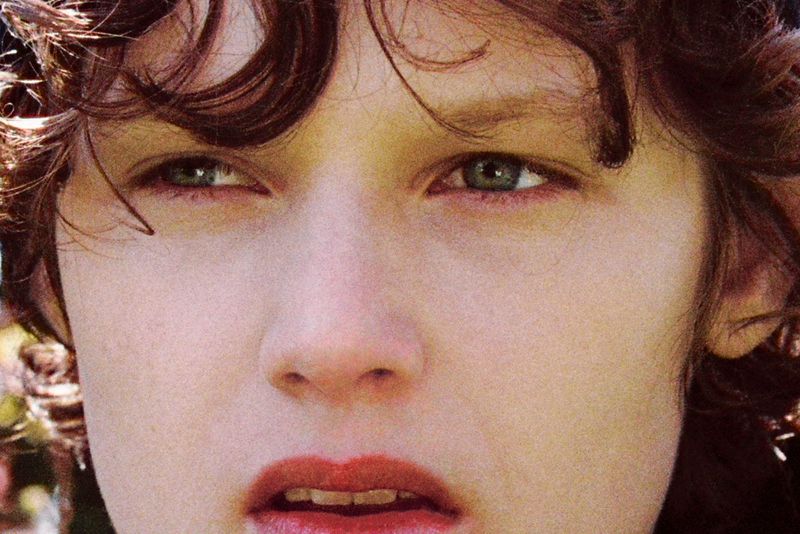 Close-up portrait of a young adult with curly hair, green eyes, and red lipstick, showcasing a beauty or fashion-related visual.