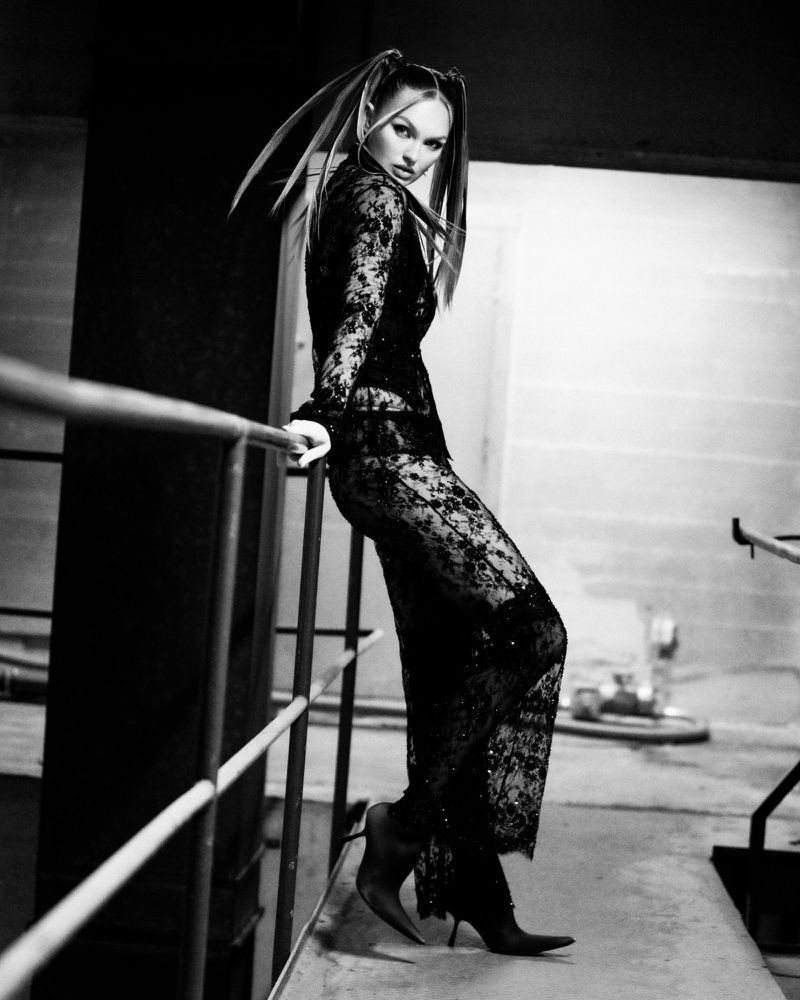 A woman poses in a black lace outfit, leaning against a metal railing in an urban setting with a concrete background and high-heeled shoes.