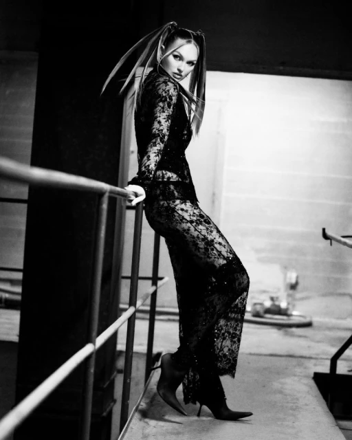 A woman poses in a black lace outfit, leaning against a metal railing in an urban setting with a concrete background and high-heeled shoes.