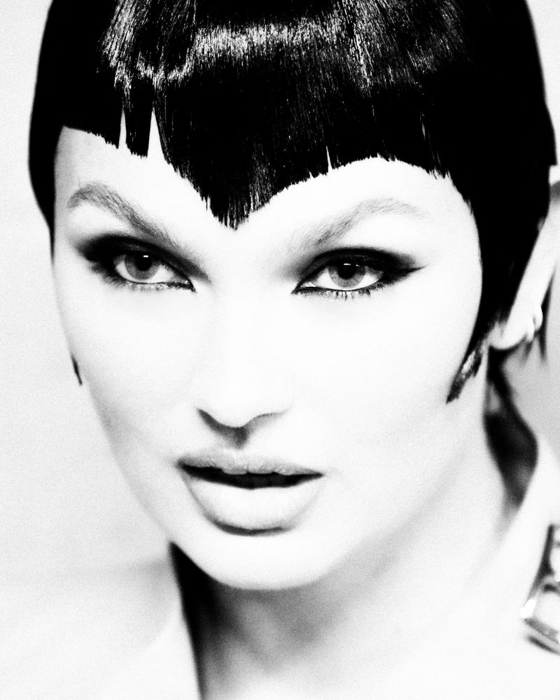 Monochrome portrait of a woman with black hair and bold makeup, featuring winged eyeliner and pale skin, posing in a light environment.
