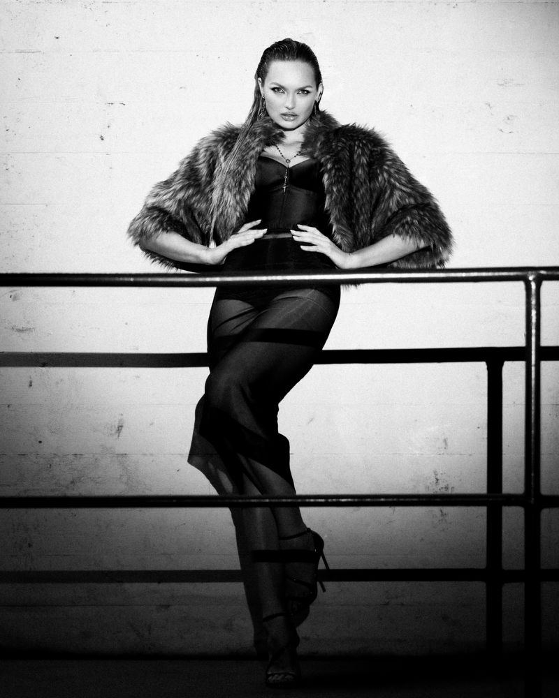 Black and white fashion portrait of a female in a fur coat, sheer skirt, and heels, posing in front of a concrete wall with metal railing.