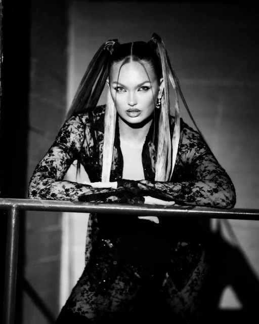 Monochrome portrait of a woman with blonde-streaked pigtails and lace top leaning on a railing, looking intensely at the camera.