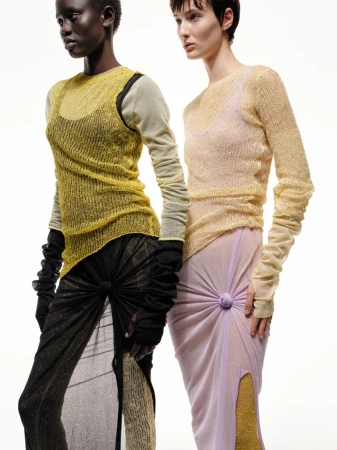 Two women pose in sheer knit tops and matching slit skirts, set against a clean white backdrop in a fashion forward editorial shot.
