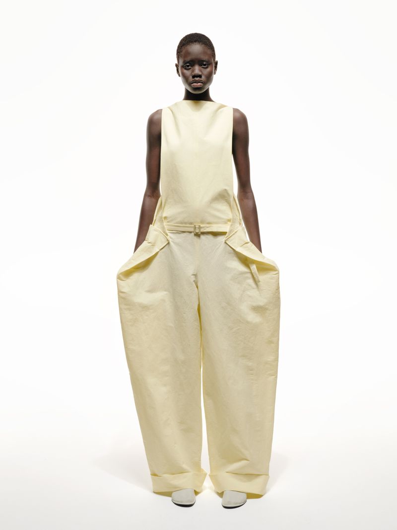 A full-length studio shot of a Black fashion model in pale yellow sleeveless top and loose-fitting trousers against a white backdrop.