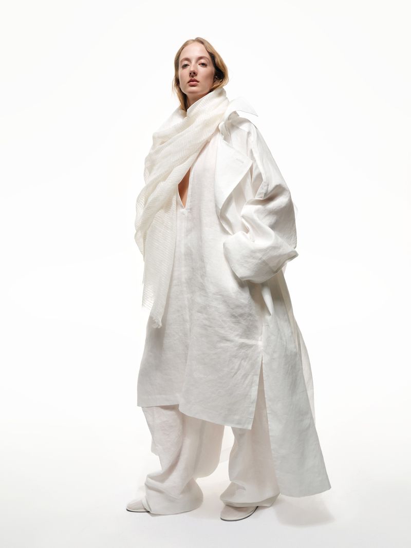 Blond woman poses in draped white linen tunic and pants against a white studio background in a bright, clean, fashion photograph.