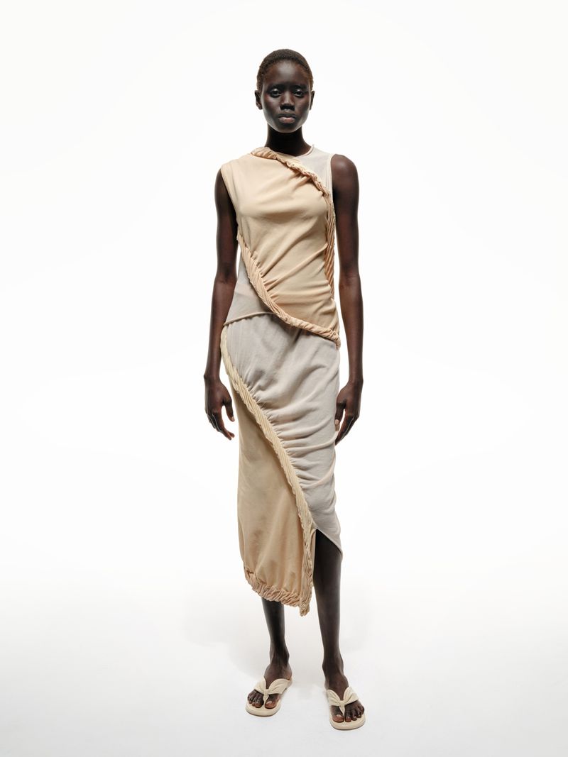 Full-length studio shot of an adult female fashion model with dark skin, posing in a draped beige dress and matching flip-flops.